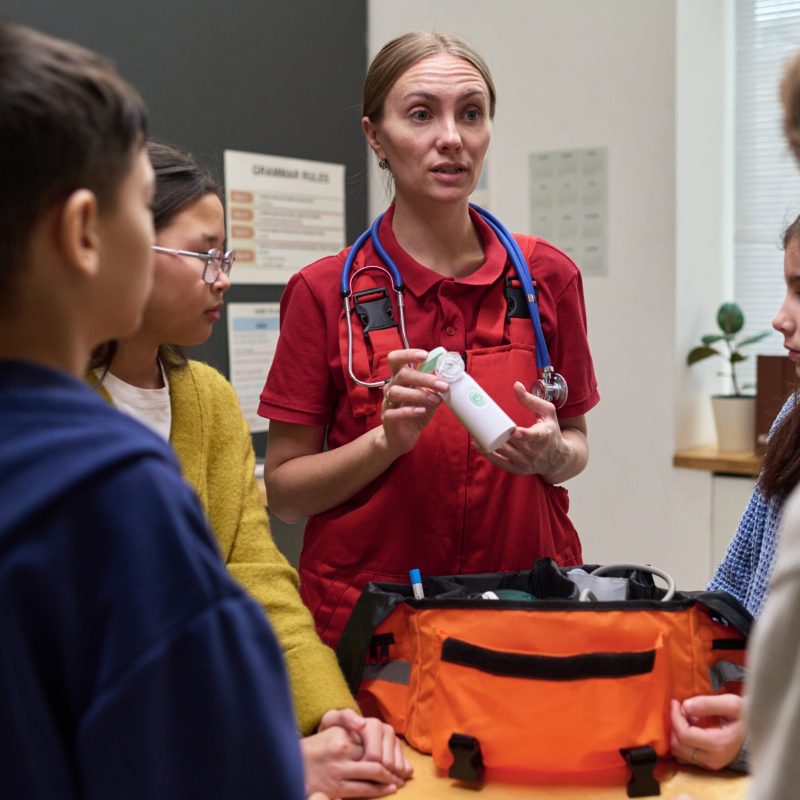 Caucasian middle aged woman paramedic demonstrating medical equipment to group of diverse children in classroom setting, children attentively listening and observing emergency kit demonstration